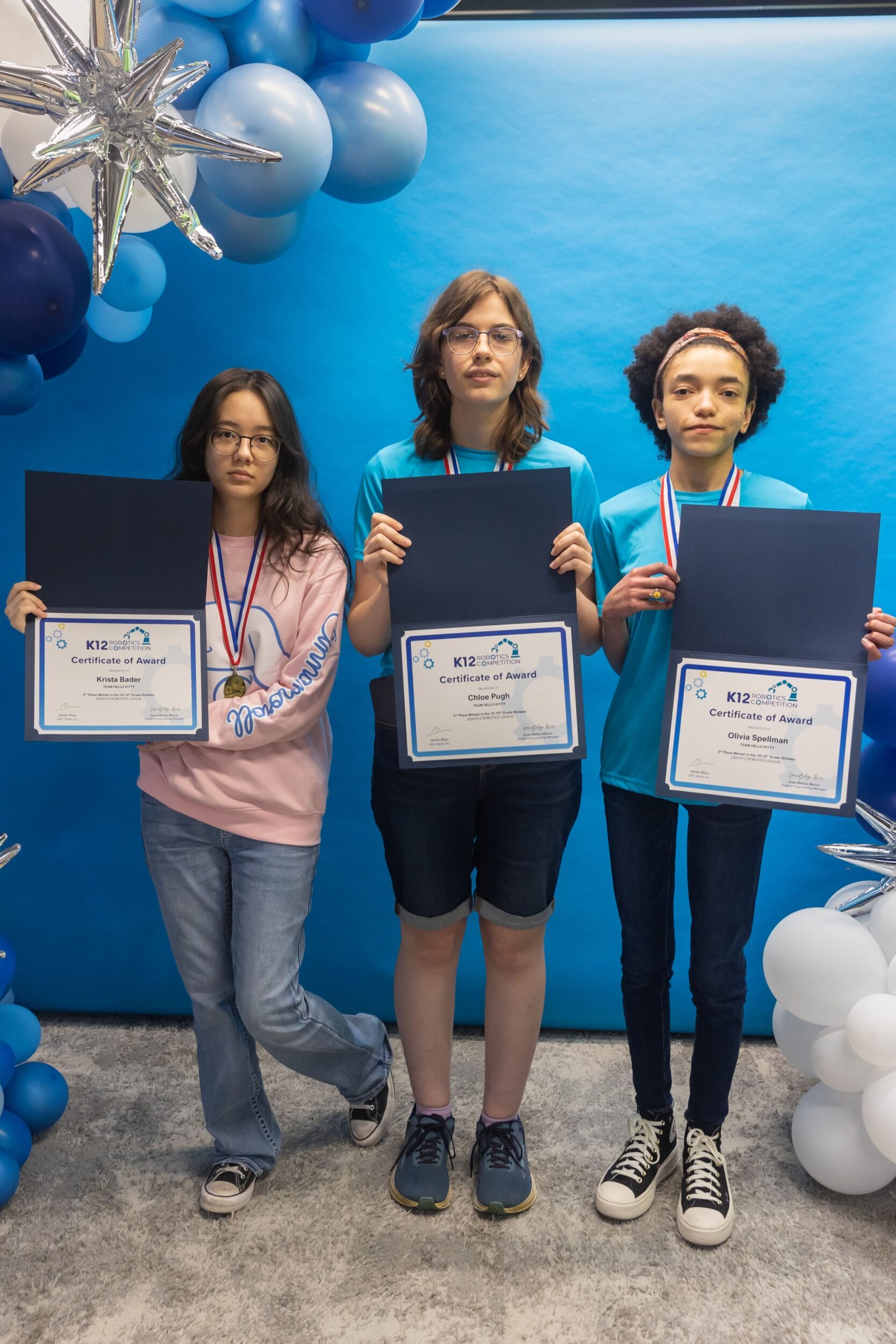 Three girls proudly display their certificates in front of a backdrop of blue balloons.